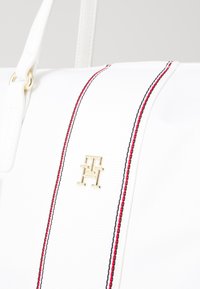 Sac à main blanc avec un logo doré, présentant une surface texturée et des accents rayés rouges et noirs sur le devant pour le détail.