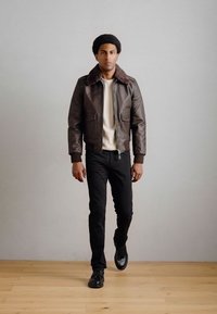 Καφέ δερμάτινο μπουφάν με γούνα στον γιακά, φορεμένο πάνω από μια κρεμ μπλούζα. Μαύροι εφαρμοστοί παντελόνια και μαύρα παπούτσια. Το μοντέλο τοποθετημένο κεντρικά.