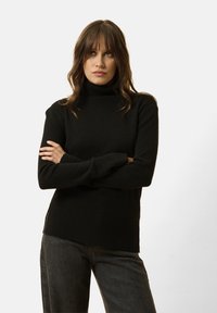 Schwarzer Rollkragenpullover aus weichem Stoff, mit geripptem Saum und Bündchen, und seitlichen Schlitzen für zusätzlichen Details. Kombiniert mit dunklen Jeans.