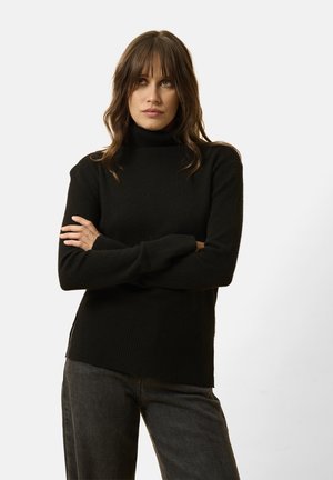 Schwarzer Rollkragenpullover aus weichem Stoff, mit geripptem Saum und Bündchen, und seitlichen Schlitzen für zusätzlichen Details. Kombiniert mit dunklen Jeans.