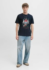 Maritime katoenen t-shirt met grafische print van een skelet in een rode hoed, gecombineerd met lichtblauwe jeans en beige schoeisel, staande tegen een witte achtergrond.