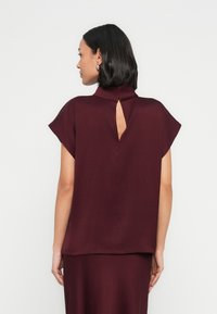 Viininpunainen paita, jossa on korkea kaulus, lyhyet hihat ja avonainen takakeskus. Sileä rakenne ja rento istuvuus. Yksinkertainen muotoilu ilman kuvioita.