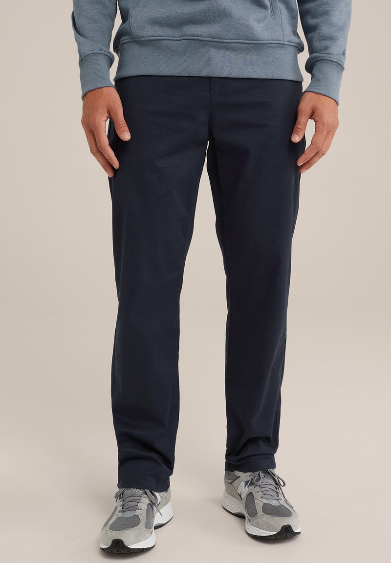 Tumši zili kokvilnas bikses ar taisnu kāju dizainu, apvienotas ar pelēkiem sporta apaviem, kas rotāti ar sietu un zamšādu.