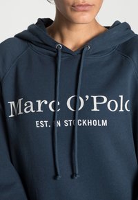 Sudadera con capucha de color azul oscuro hecha de una tela suave. Presenta un cordón en la capucha y un texto impreso en blanco que dice "Marc O'Polo EST. EN ESTOCOLMO."