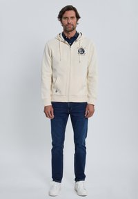 Sudadera con capucha de cremallera beige con bolsillos tipo canguro, puños acanalados y capucha con cordón, combinada con vaqueros azul oscuro y zapatillas blancas.