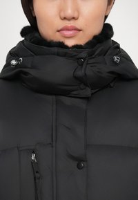 Jachetă puffer neagră cu guler înalt, având căptușeală din blană sintetică, accesorii argintii și negre, precum și închideri cu fermoar și nasturi.