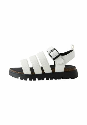 Weiße Ledersandale mit drei Riemen, schwarzer Schnalle, Korkfußbett und dicker schwarzer profilierter Sohle, Seitenansicht vor weißem Hintergrund.
