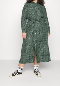Robe verte cintrée avec un motif de feuilles, manches longues, taille ceinturée et fente à l'avant, associée à des baskets noires et des chaussettes rayées.