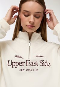 Kurtka z zamkiem w kolorze kremowym z wyszywanym napisem "Upper East Side" w burgundzie, z wysokim kołnierzem i ściągaczami przy mankietach.