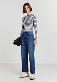 Grau-weißer gestreifter, gerippter Langarm-Top, hochtaillierte blaue gerade Jeans, mit weißen und beigen Sneakers, hält eine schwarze Tasche.