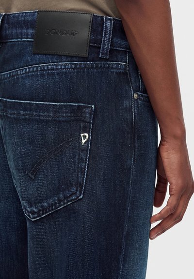 Poche arrière d'un jean en denim bleu foncé avec un petit logo métallique, un patch de marque en cuir noir et la main d'une personne reposant à côté de la hanche.