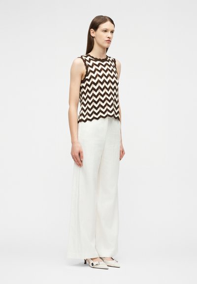 Γυναίκα όρθια, φορώντας μπλουζάκι χωρίς μανίκια με καφέ και λευκό ζιγκ-ζαγκ σχέδιο, φαρδιά λευκή παντελόνα και λευκά παπούτσια με χαμηλό τακούνι και αγκράφες.