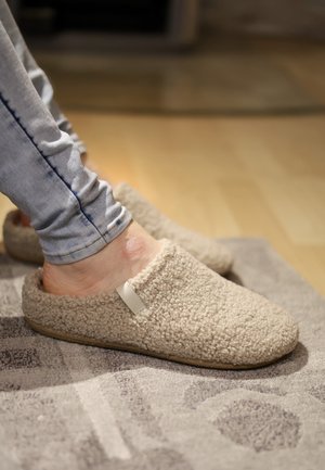 Füße in beigen, flauschigen Hausschuhen auf einem grau gemusterten Teppich, dazu hellblaue Jeans sichtbar.