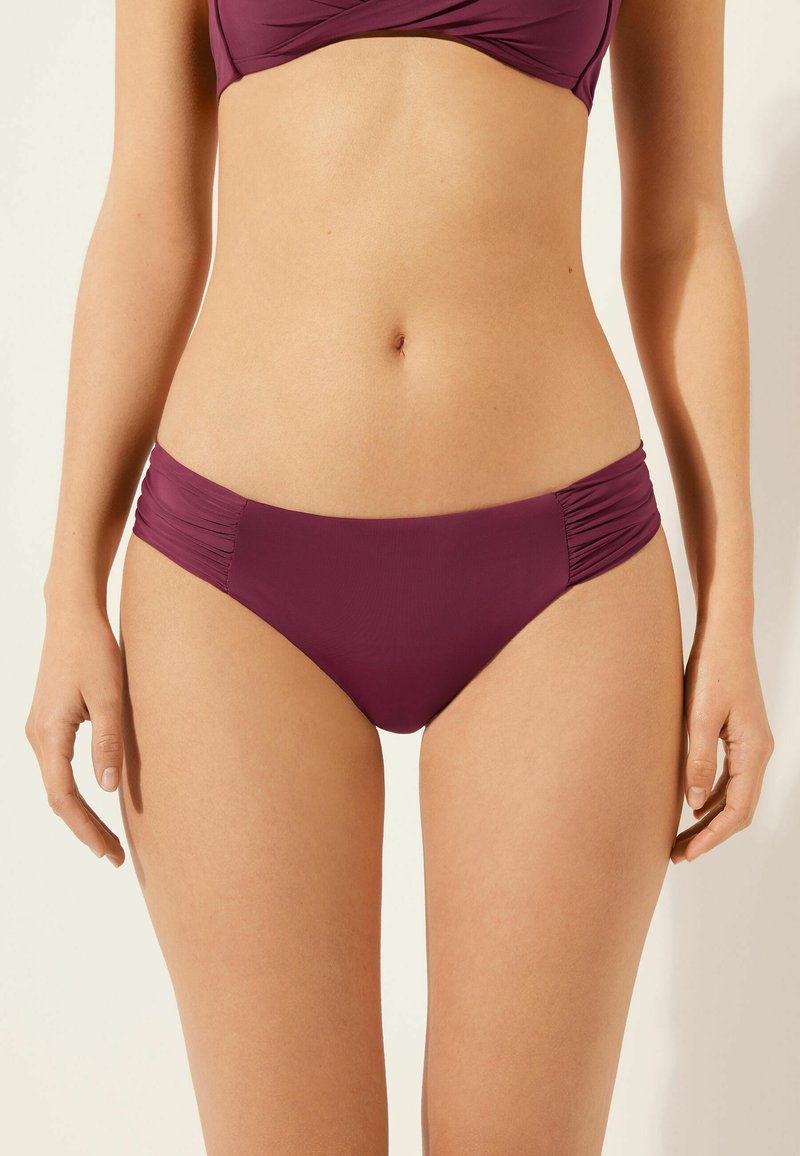 Bas de bikini en bourgogne foncé avec des détails froncés sur les côtés, fabriqué à partir d'un tissu lisse et extensible, présentant une coupe modérée et un design sans couture.