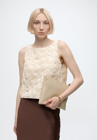 Femme aux cheveux blonds courts porte un haut sans manches beige texturé, une jupe marron et un bracelet doré tout en tenant une pochette beige.