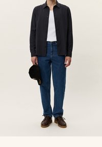 Dunkles Hemd mit Knöpfen über einem weißen Tanktop, kombiniert mit blauen Jeans und braunen Schnürschuhen. Eine schwarze Baseballkappe in der Hand.