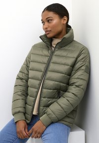 Olijfgroene pufferjack met een hoge kraag, een gewatteerd ontwerp, zijzakken met rits en geribbelde cuffs, gecombineerd met een beige trui en blauwe jeans.