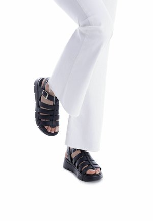 Sandalias negras con tiras y suela acolchada, que presentan múltiples tiras horizontales y una hebilla lateral, llevadas con pantalones blancos de pierna ancha.