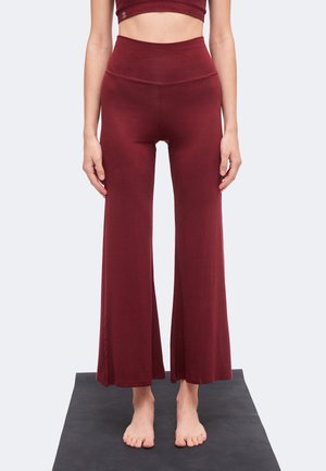 Femme debout pieds nus sur un tapis de yoga, portant un pantalon bordeaux taille haute à jambes larges et un crop top assorti, les bras le long du corps.