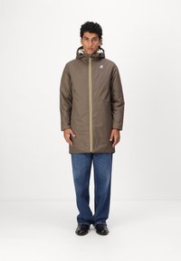 Brown waterproof coat with hood and yellow zipper, featuring a fleece-lined collar. Worn over blue denim pants and black shoes.