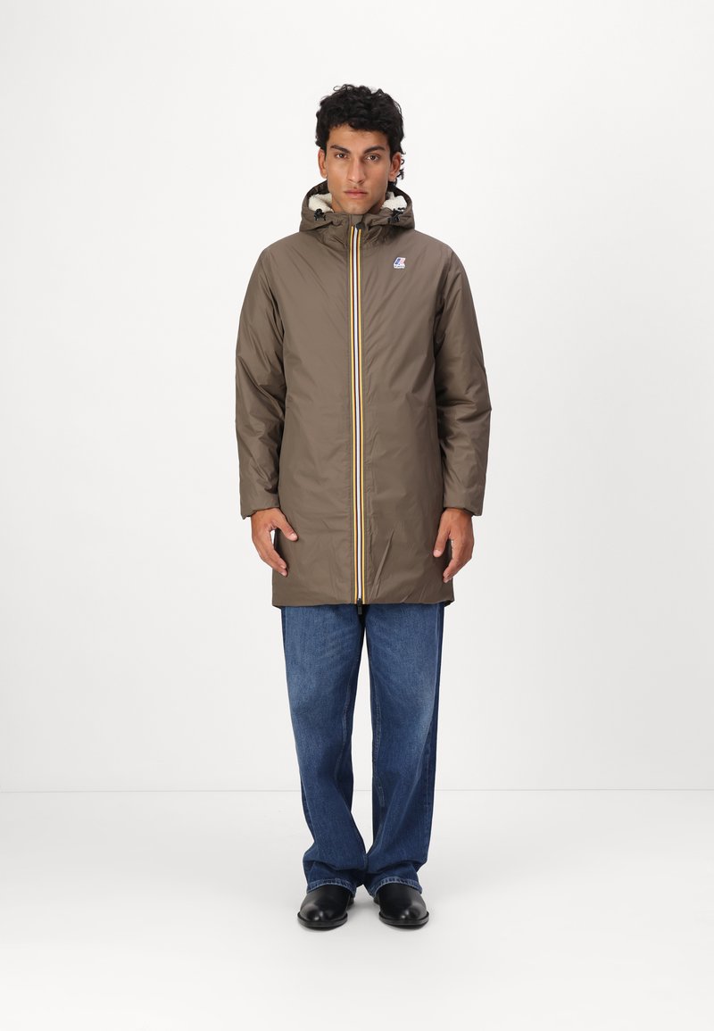 Brown waterproof coat with hood and yellow zipper, featuring a fleece-lined collar. Worn over blue denim pants and black shoes.