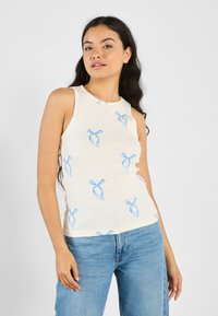Ribbed beige tank top with blue bow pattern, featuring a round neckline and sleeveless design. Paired with light blue jeans.