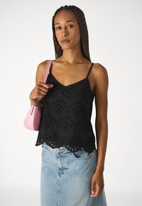 Top à bretelles noir brodé avec un ourlet festonné, mettant en valeur des découpes florales ; associé à une jupe en denim clair et à un petit sac à main rose.