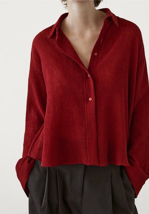 Chemise rouge texturée avec col, manches longues et coupe ample, portée avec un pantalon foncé. Tissu doux avec un motif en relief subtil.