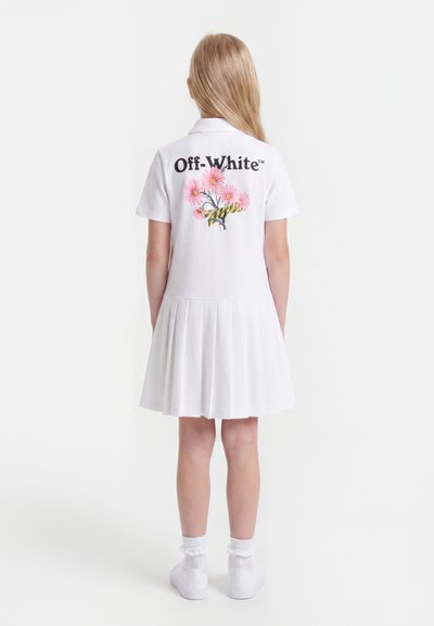 Κορίτσι που φοράει λευκό πλισέ φόρεμα με το κείμενο «Off-White» και γραφικά με ροζ λουλούδια στην πλάτη, συνδυασμένο με λευκές κάλτσες και αθλητικά παπούτσια.