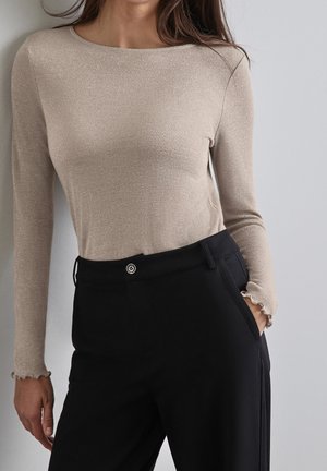 Frau trägt ein beiges, langärmliges, eng anliegendes Oberteil mit dezentem Schimmer und schwarze, hochtaillierte Hosen, eine Hand in der Tasche.