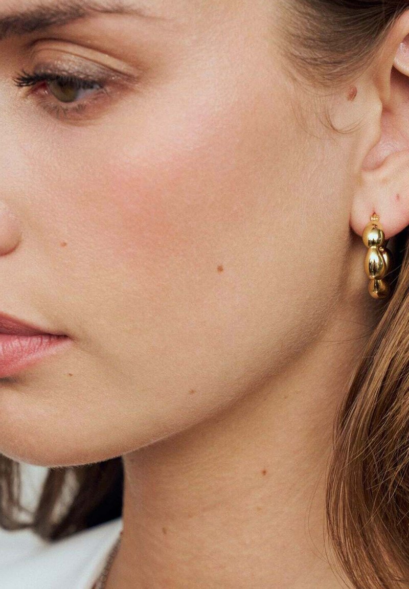 Guld oval hoopörhängen med en slät, reflekterande yta. Har en staplad pärldesign som visar upp en polerad textur och subtil glans.