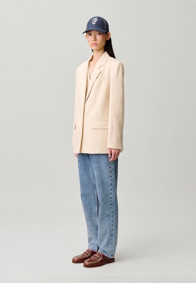 Blazer beige a righe sottili, jeans denim blu dal taglio morbido, mocassini marroni e cappellino da baseball blu navy con logo bianco. Silhouette semplice e moderna.