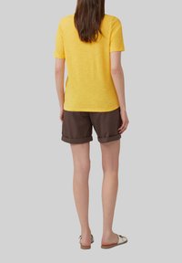 Chemise jaune à manches courtes, texture douce, associée à un short brun roulé. La tenue est complétée par des sandales à enfiler de couleur claire.