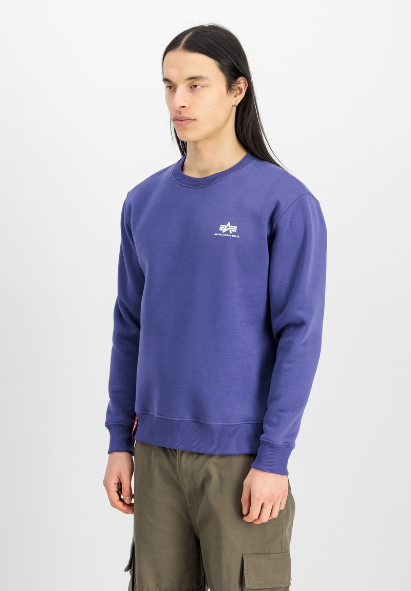 Lila crewneck pulóver, 100% pamutból készült, kis fehér logóval a mellrészén, bordázott mandzsettákkal és laza szabással. Olíva zöld cargo nadrággal viselve.