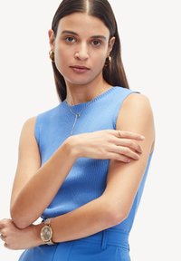 Gestreepte blauwe mouwloze top gecombineerd met een gouden horloge en sieraden; focus op textuur en aansluitend ontwerp, armen gekruist voor een nette presentatie.