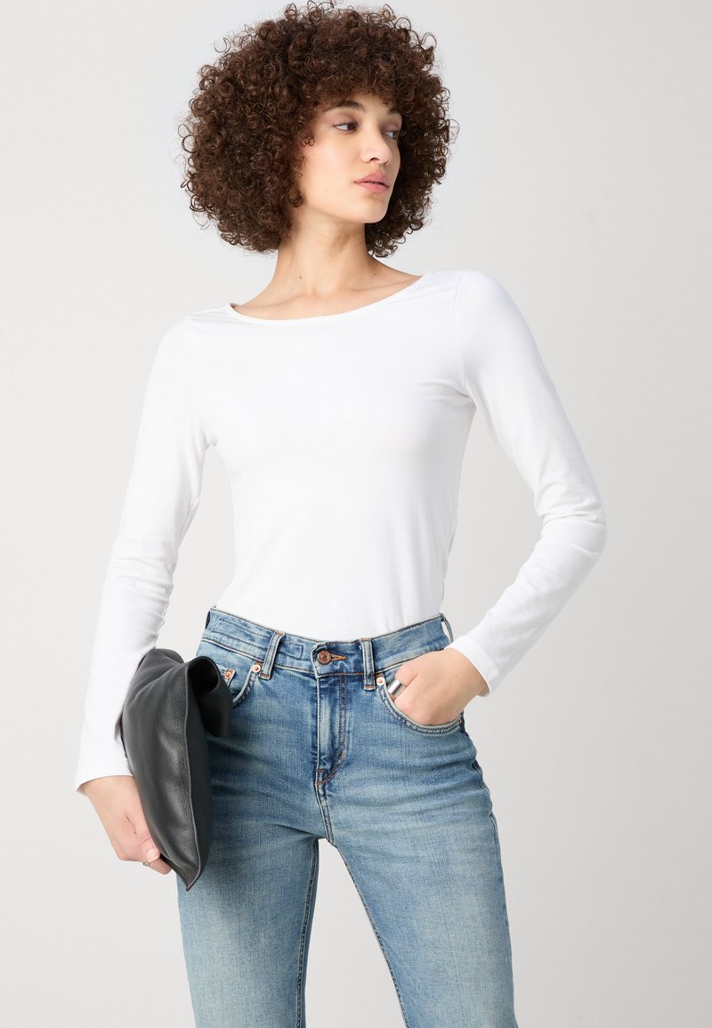 T-shirt blanc à manches longues en tissu extensible associé à un jeans taille haute bleu clair. Le modèle tient une pochette en cuir noir.