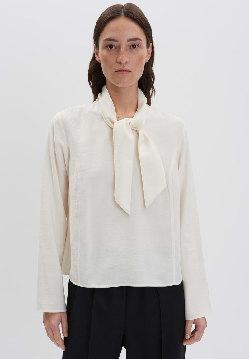 Blusa color crema con colletto a fiocco, maniche lunghe e texture liscia; abbinata a pantaloni neri larghi.
