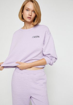 Lila texturerad sweatshirt med långa ärmar och rundad halsringning, med en broderad "New York" detalj. Matchas med tillhörande byxor.