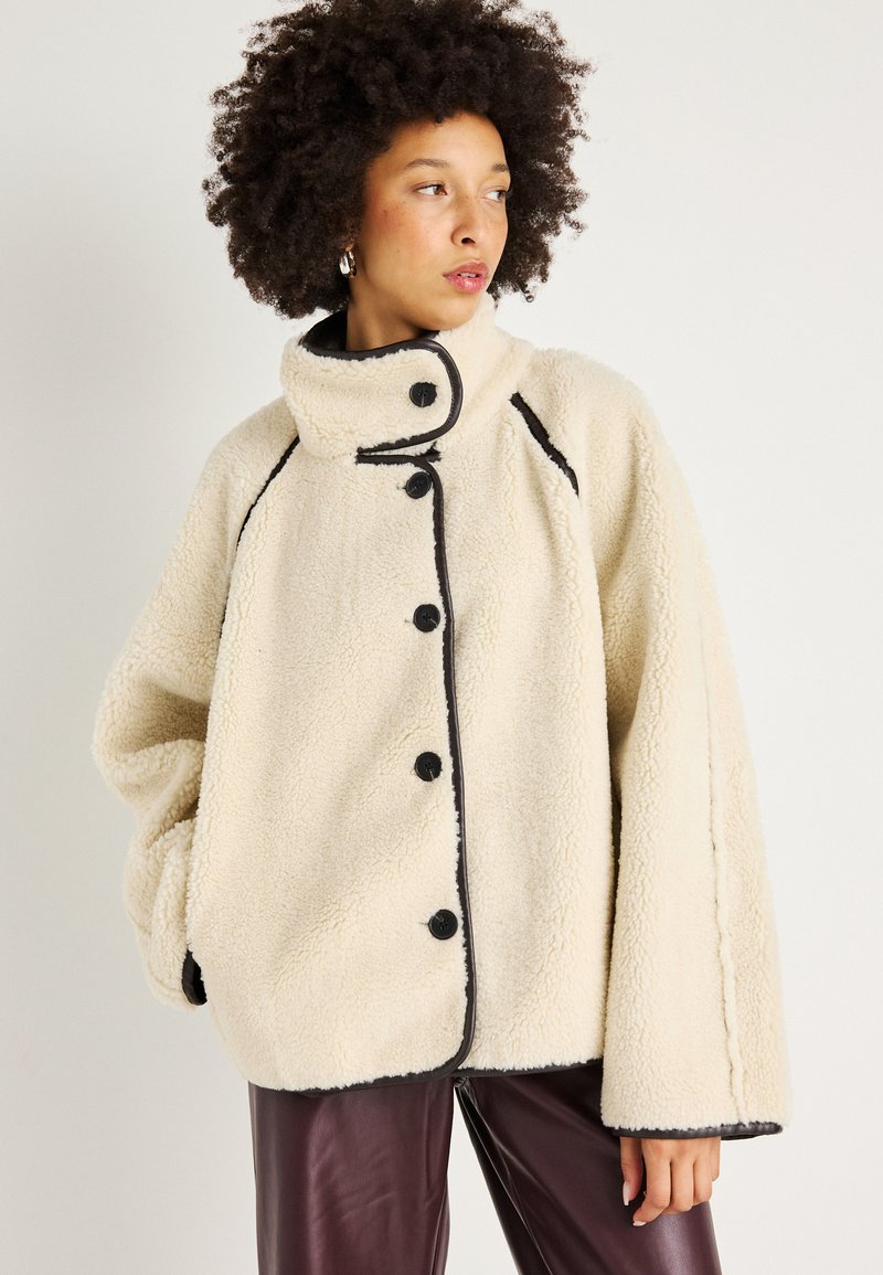 Crème-kleurige fleece jack met zwarte bies, hoge kraag en grote knopen. Heeft een losse, oversized pasvorm en een zachte structuur.