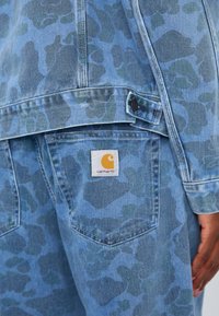 Denimjakke med et blåt camouflage-mønster, prydet med et Carhartt-logo patch. Stoffet har en tekstureret overflade og synlige sømdetaljer.