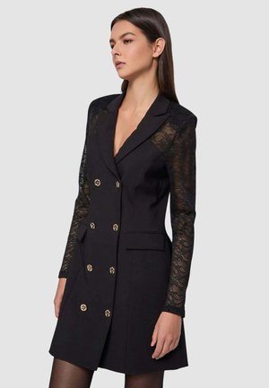 Donna che indossa un abito blazer doppiopetto nero con bottoni dorati e maniche lunghe in pizzo trasparente, in piedi davanti a uno sfondo neutro.