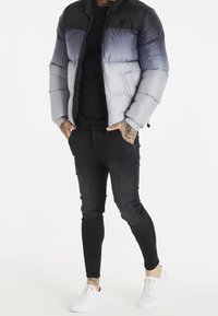 Mu muž v šedém gradientním prošívaném kabátu, černých skinny džínách a bílých teniskách, stojící s rukama v kapsách na bílém pozadí.