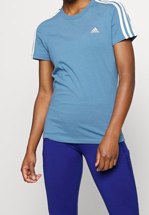 Osoba ubrana w niebieski t-shirt Adidas z białymi paskami na rękawach i ciemnoniebieskie legginsy, stoi na jednolitym tle.