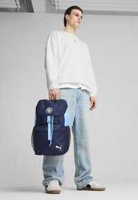 Zaino blu navy e azzurro chiaro con chiusura a coulisse, tasche laterali in rete, logo Puma e distintivo circolare sulla parte anteriore.