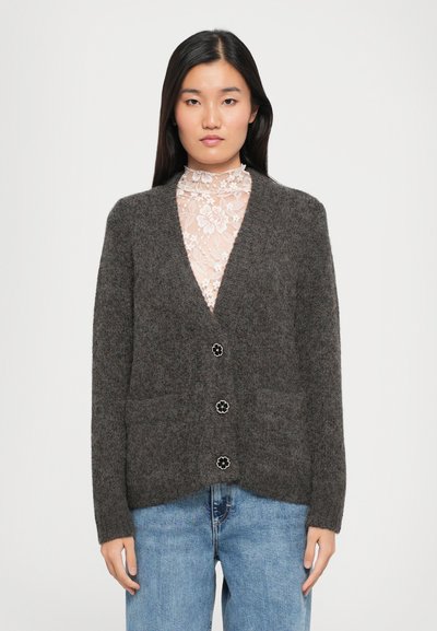 Cardigan grigio a maglia con scollatura a V, due tasche frontali e bottoni con design floreale. Indossato sopra un top di pizzo, abbinato a jeans azzurri chiari.