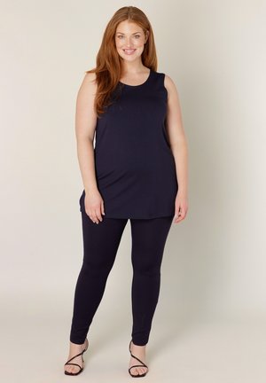 Kromme vrouw met lang rood haar, gekleed in een mouwloze zwarte tuniek, zwarte legging en zwarte sandalen met bandjes, staand en glimlachend.