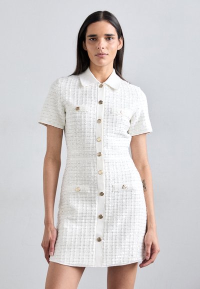 Robe blanche à manches courtes avec un tissu texturé, un design à boutons sur le devant et des accents en métal doré sur les poches.