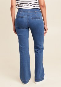 Bootcut jeans in middelblauw denim met een gladde textuur, twee achterzakken en lichte vervaging aan de zoom. Gecombineerd met witte sneakers.