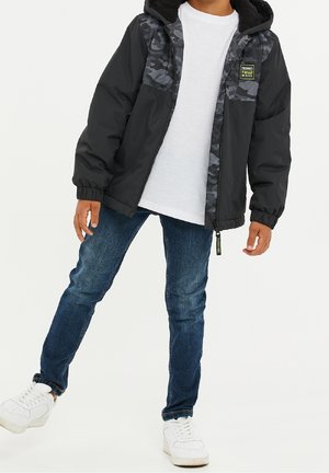 Kind, der eine schwarze Jacke mit Camouflage-Muster und Kapuze trägt, ein weißes T-Shirt, eine blaue Jeans und weiße Turnschuhe, steht mit einem Fuß leicht angehoben.