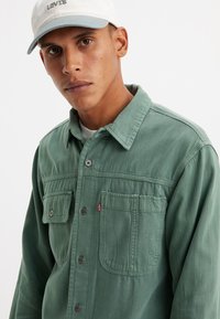 Camisa de algodón verde con dos bolsillos frontales, cierre de botones y detalles de costura visibles; combinada con una gorra de color claro.